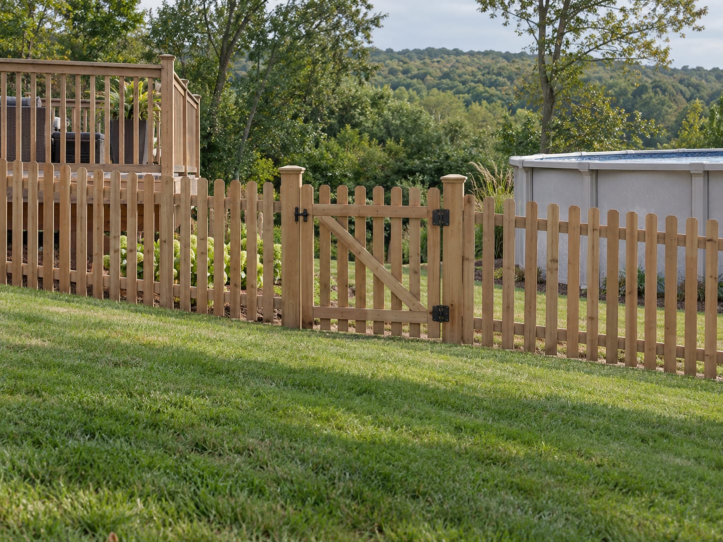 A tall cedar picket fence with a closed gate stands between a low wooden deck and an above-ground pool on a gently sloped backyard.