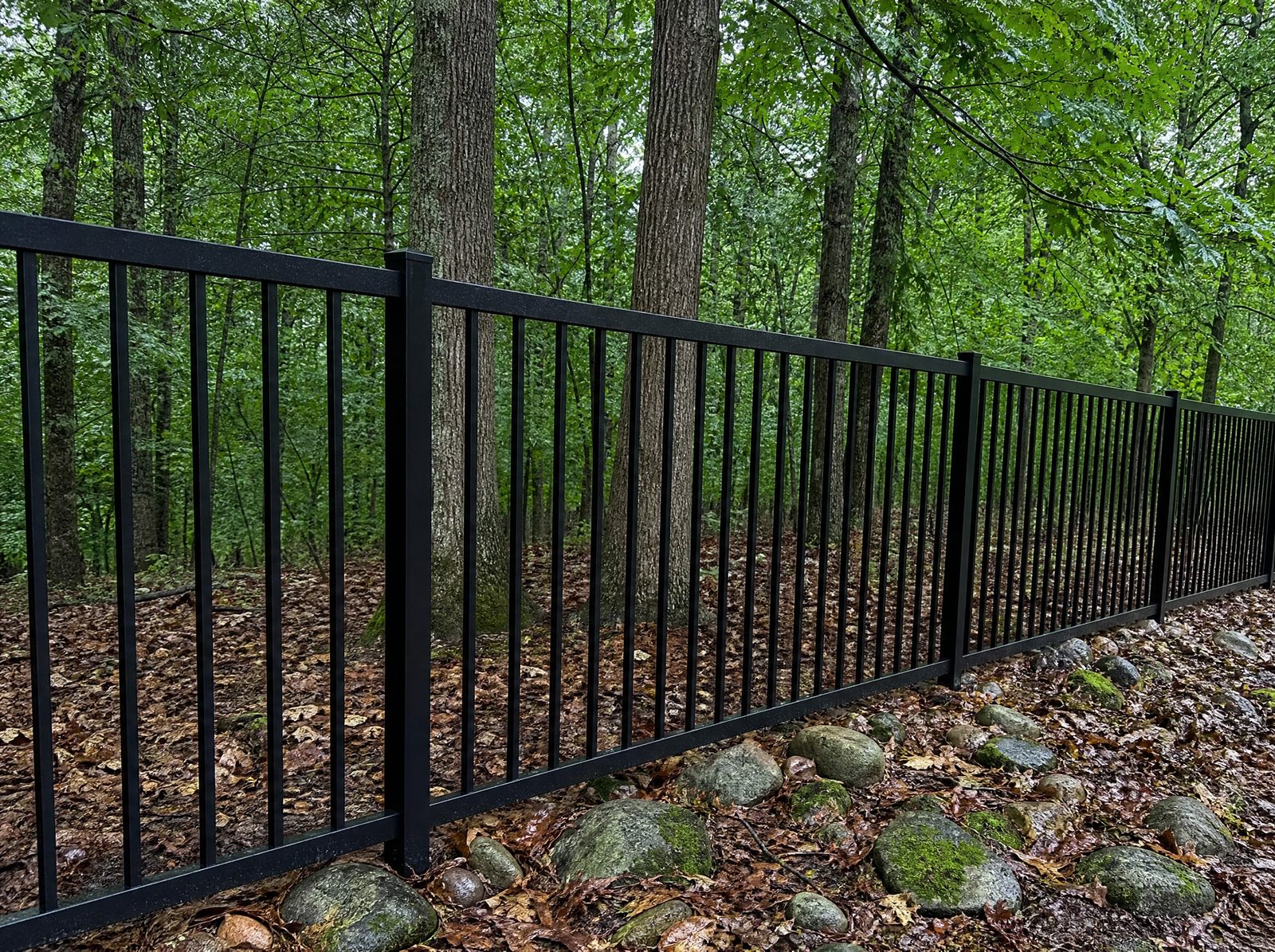 Four foot black flat-top aluminum fence holding a level top line along a wooded rural rear property boundary over uneven ground