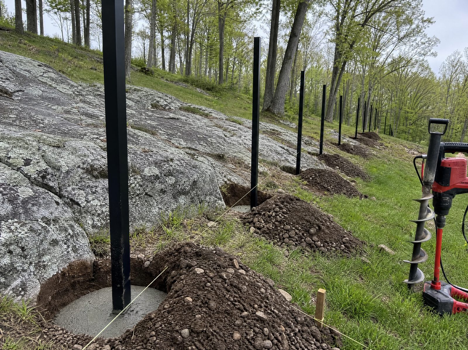Black aluminum fence posts set plumb in freshly augered holes stepping down a rocky sloping rural lot with exposed ledge and no panels hung yet