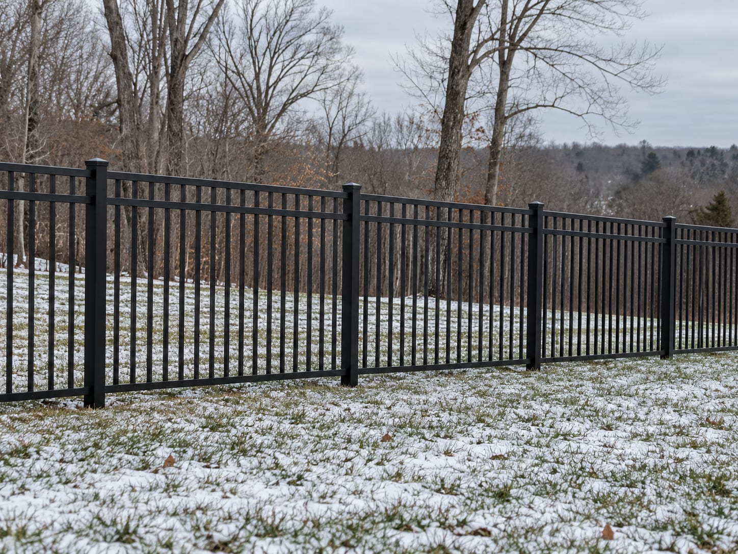 A black aluminum fence runs along a lawn with a light dusting of snow on the ground and bare trees beyond in western Connecticut.