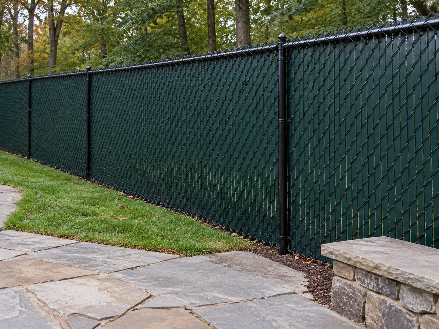 A black vinyl-coated chain link fence with green privacy slats woven through the mesh runs along the edge of a patio and lawn in western Connecticut.