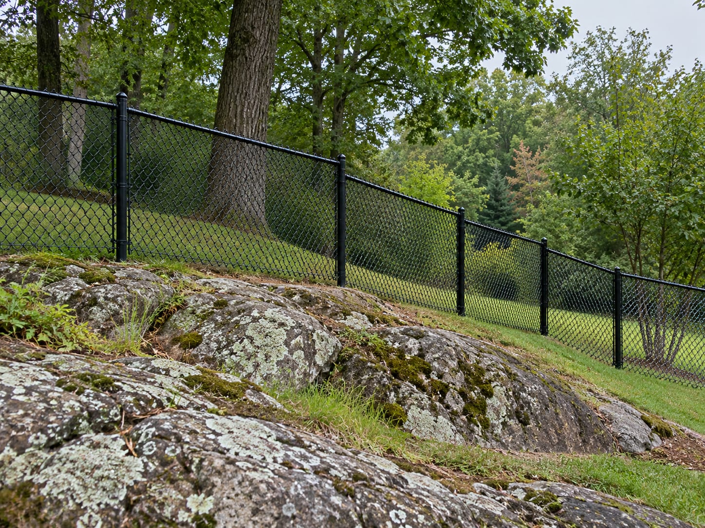 A black vinyl-coated chain link fence steps down a gentle slope beside an exposed ledge outcrop in a western Connecticut backyard.