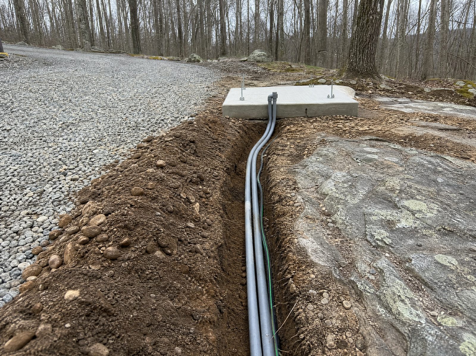 Grey electrical conduit and low-voltage wiring lying in an open trench along the edge of a Sherman gravel driveway running toward a poured concrete gate operator foundation pad