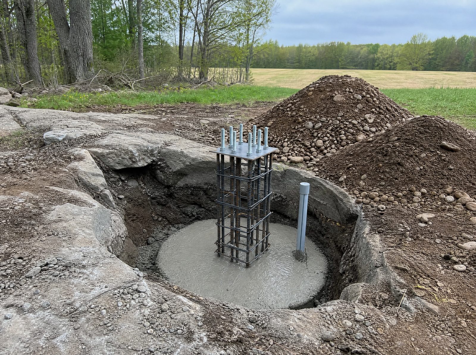 Deep concrete gate-post footing being set below frost in rocky Southbury ground with a rebar cage and anchor bolts standing proud of the wet concrete and no gate leaf hung yet