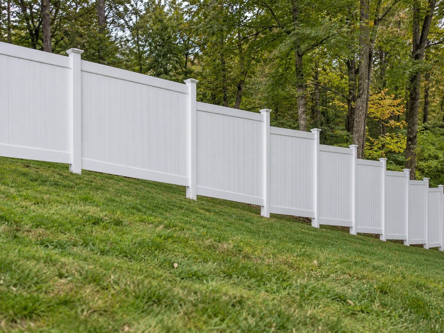 A white vinyl privacy fence stepped down a gentle grassy slope in level sections, with the ground falling away beneath each panel, in western Connecticut.