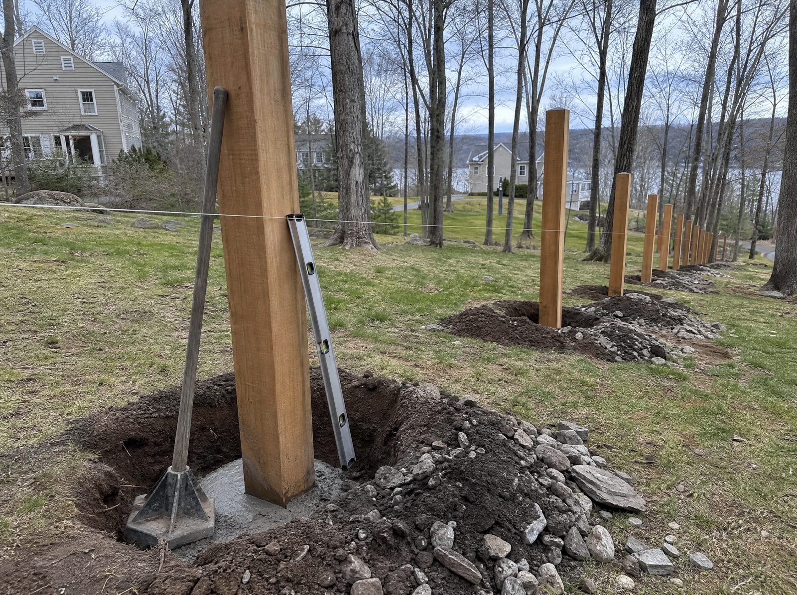 Freshly set wood fence posts standing plumb in deep holes down a sloped rocky lot with a string line