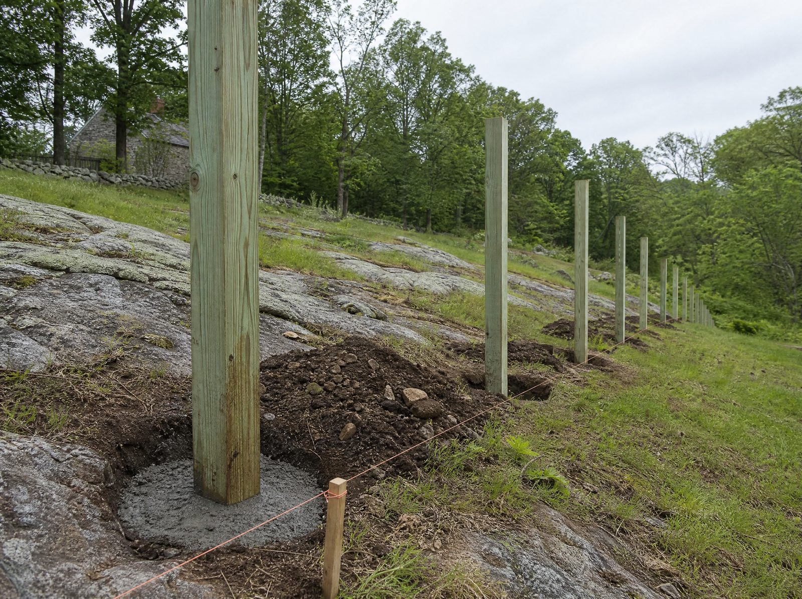 Pressure-treated fence posts set plumb in freshly augered deep holes along a rocky sloping rural lot with exposed ledge