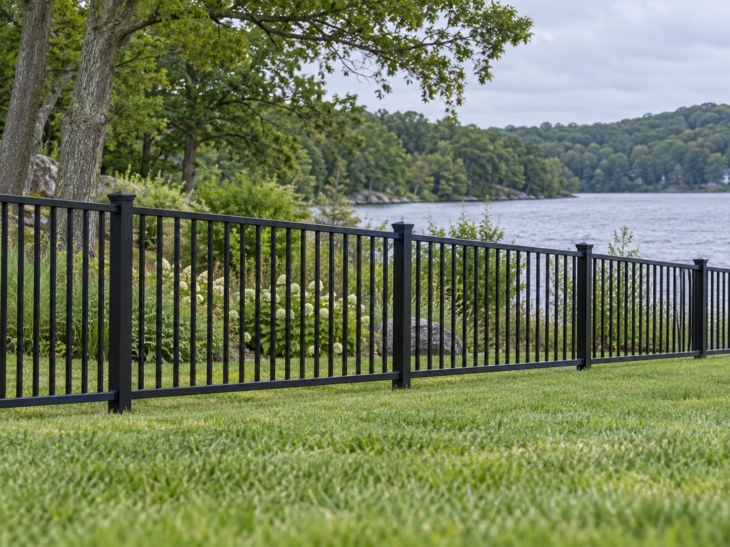 A black aluminum fence with a small open gap beneath the bottom rail, running along a lawn that slopes toward a quiet lake shore with native shrubs in western Connecticut.