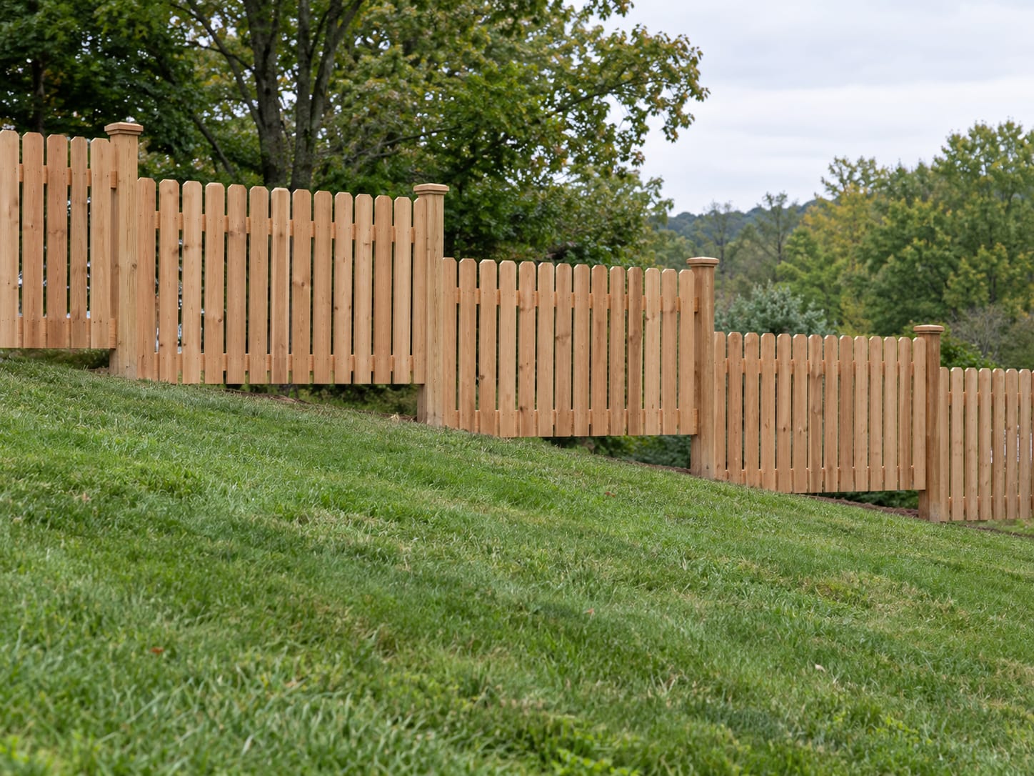 A stepped cedar picket fence on a sloping lawn with level sections and a triangular open gap beneath the downhill end of each section, in western Connecticut.