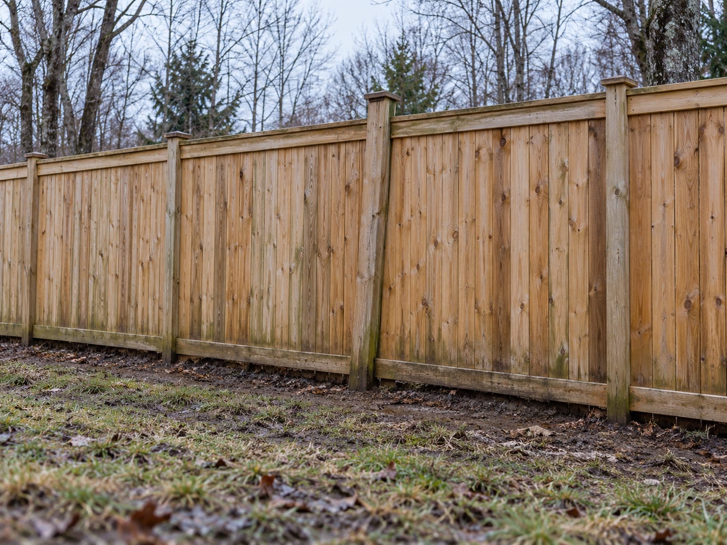 A wood privacy fence on damp early-spring ground where one post has lifted slightly and leans out of line with its neighbors.