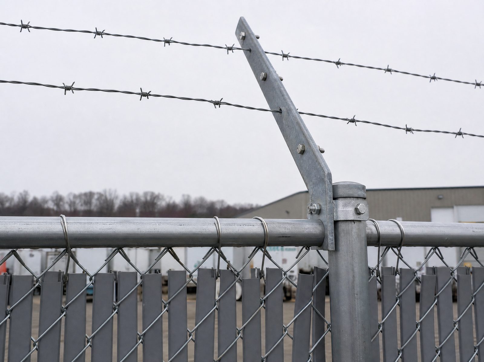 Close honest view of a three-strand barbed-wire outrigger arm angled outward from the top rail of a tall galvanized industrial chain-link fence above the slatted mesh