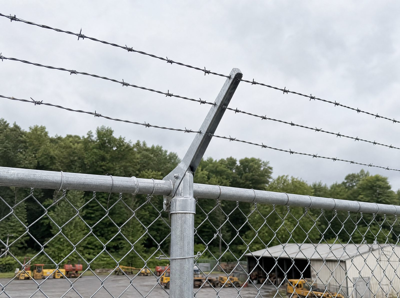 Close honest view of a three-strand barbed-wire outrigger arm angled outward from the top rail of a tall galvanized industrial open-mesh chain-link fence