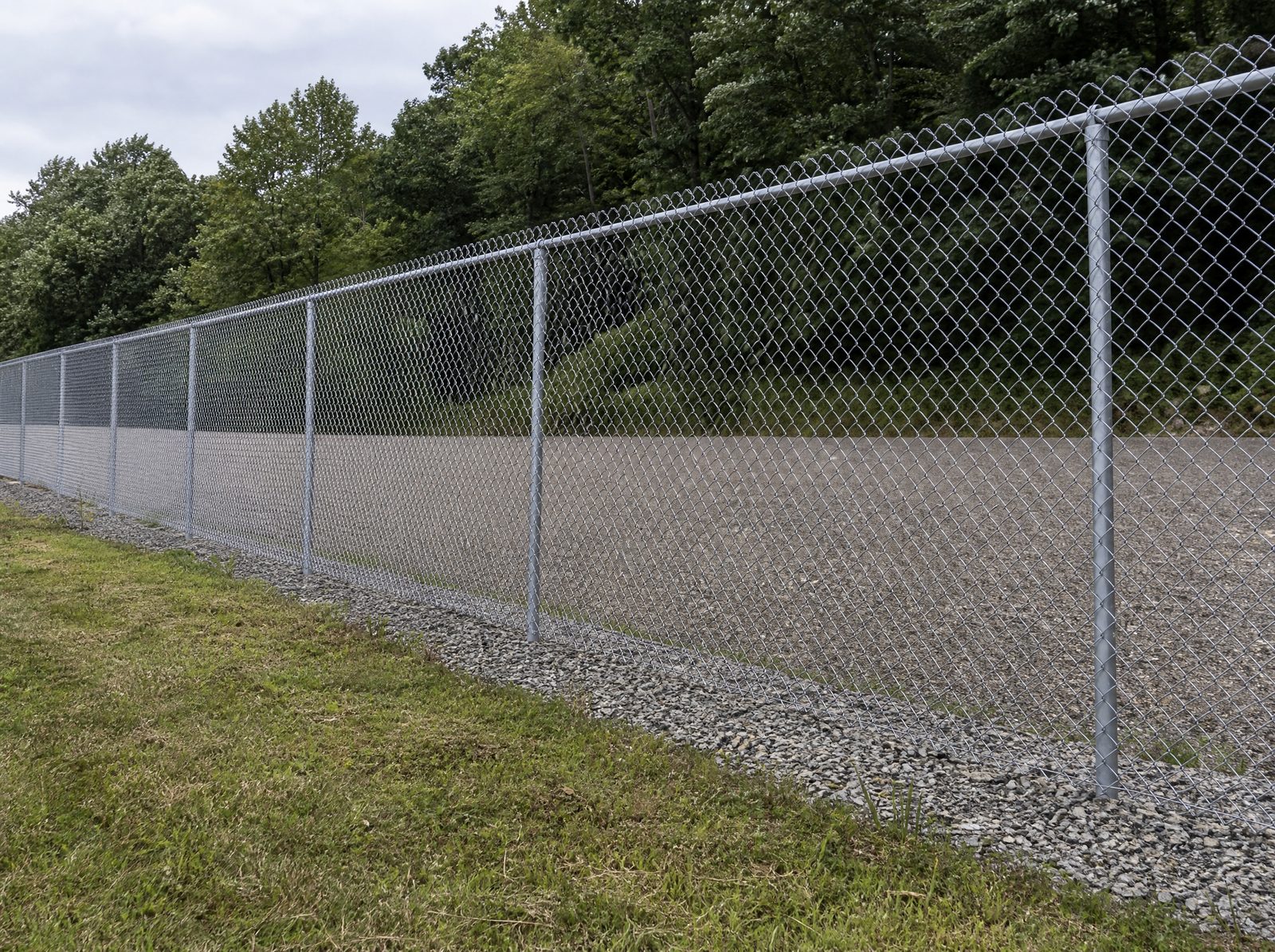 Eight foot galvanized chain link perimeter fence running along the edge of an equipment yard with a wooded ridge rising behind it