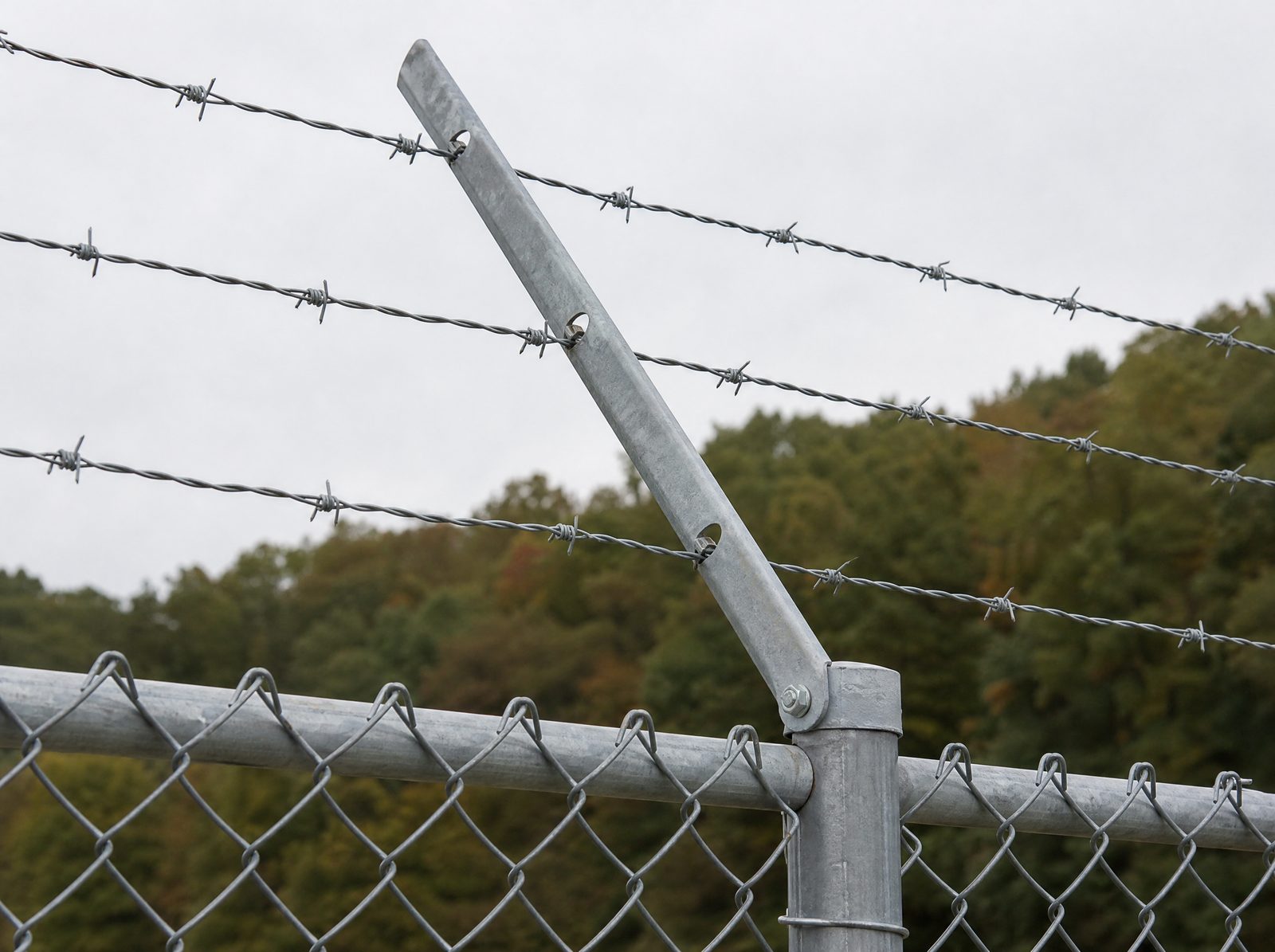 Close view of a three strand barbed wire outrigger arm angled outward from the top rail of a tall galvanized industrial chain link fence