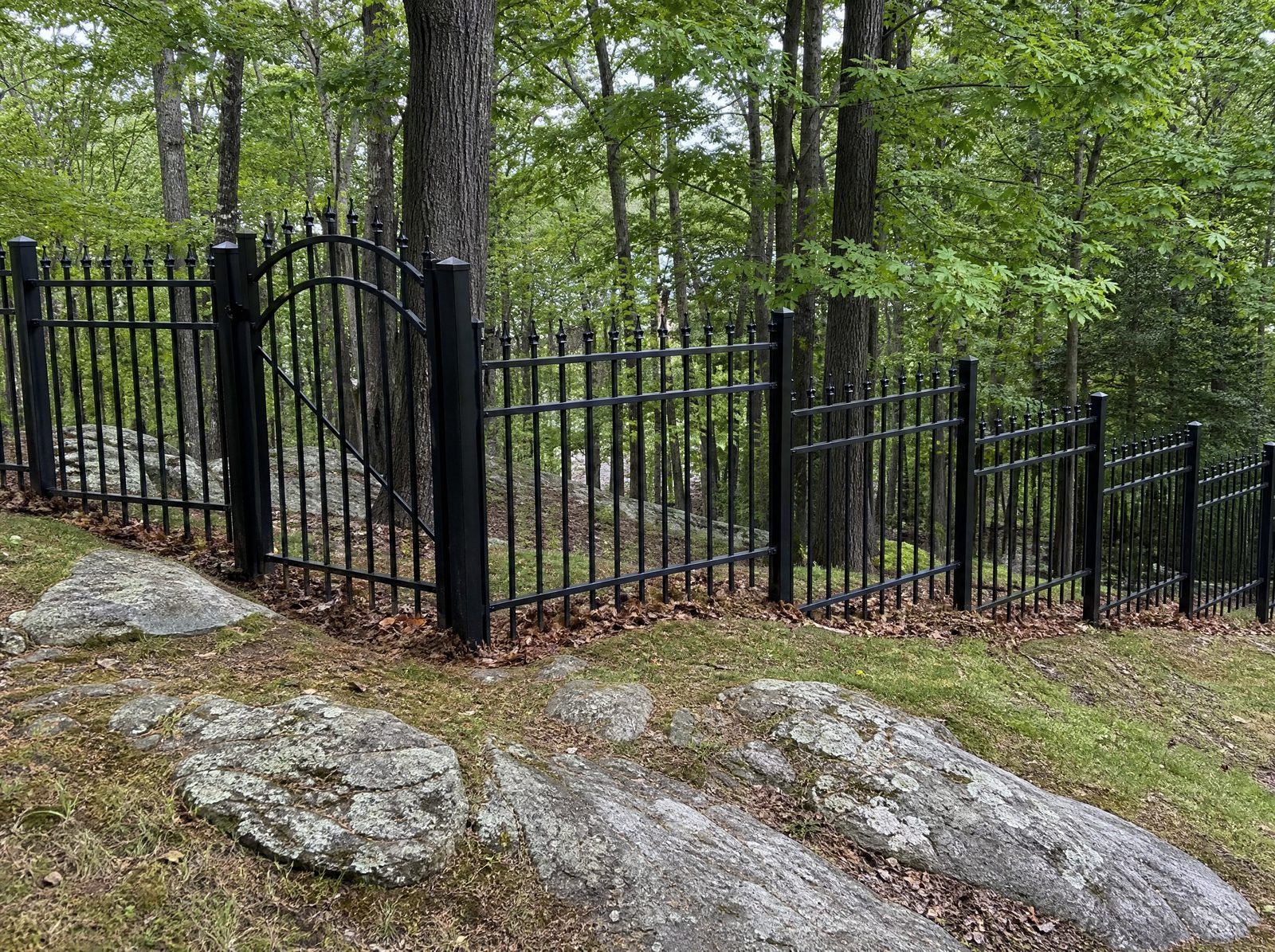 Four foot black ornamental metal fence with finials stepping down a sloped wooded yard in level panels with an arched entrance gate on the flattest span