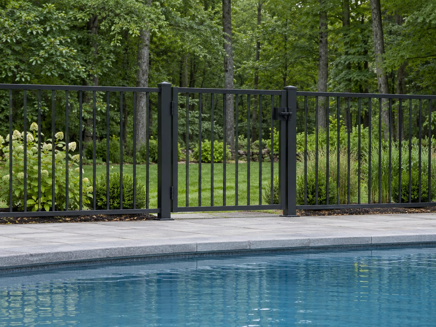 A black aluminum fence with a closed self-latching gate surrounds an in-ground pool on a level backyard, with hardwood trees beyond the fence.