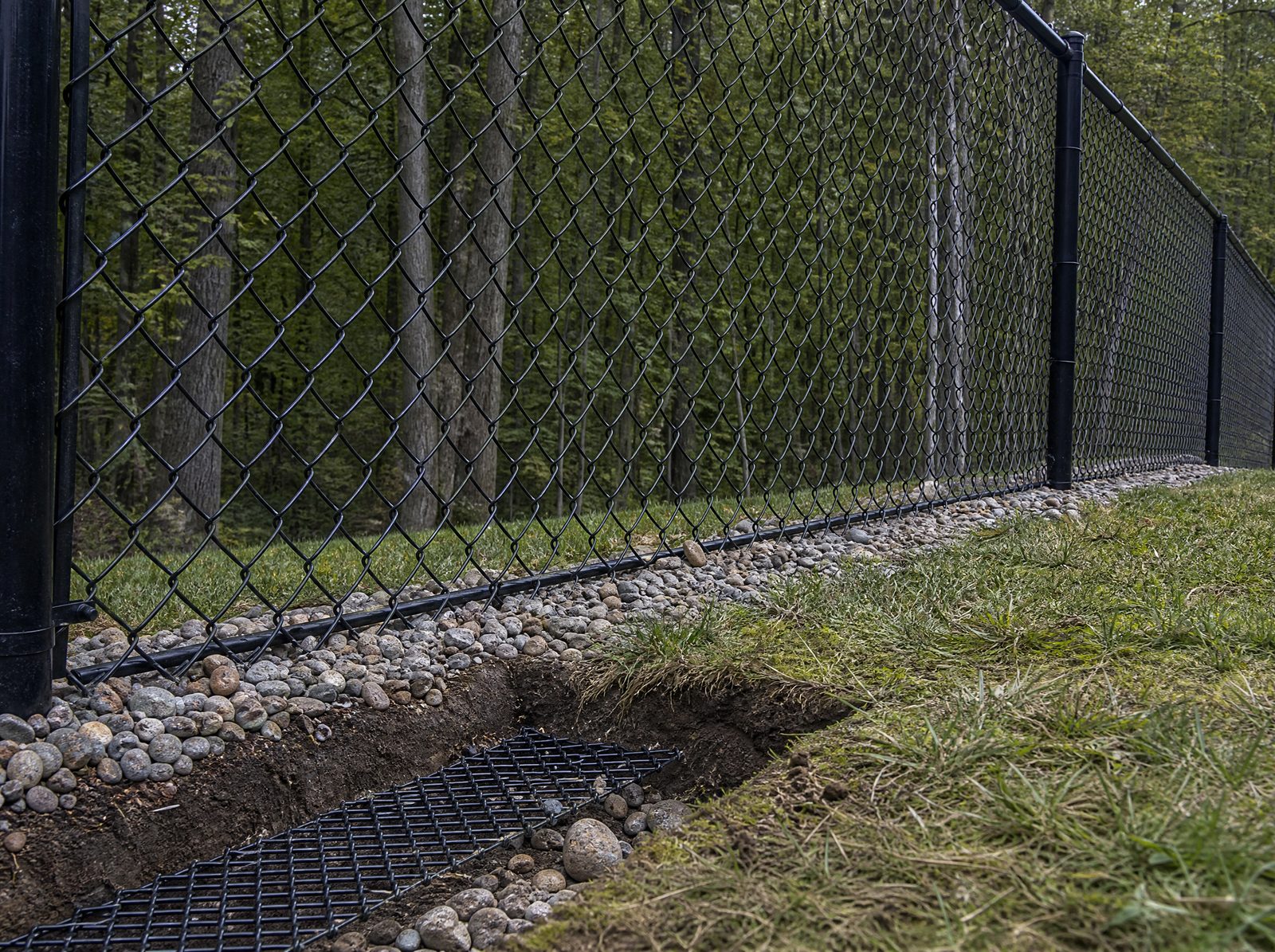 Base of a black chain link dog fence running along a sloped backyard with the bottom edge held tight to the grade over a buried L shaped footer, no dog present