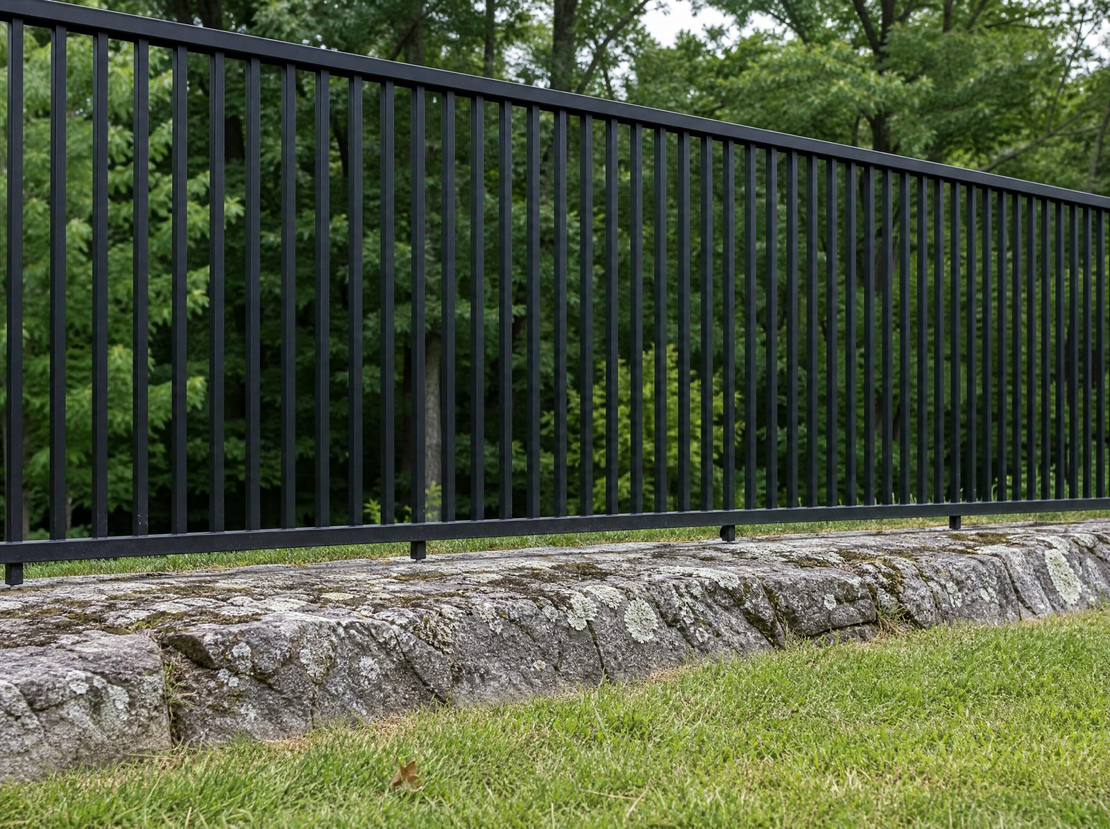 Straight stretch of black aluminum pool barrier close enough to read the tight vertical picket spacing and the small even ground clearance under the bottom rail running over grey ledge