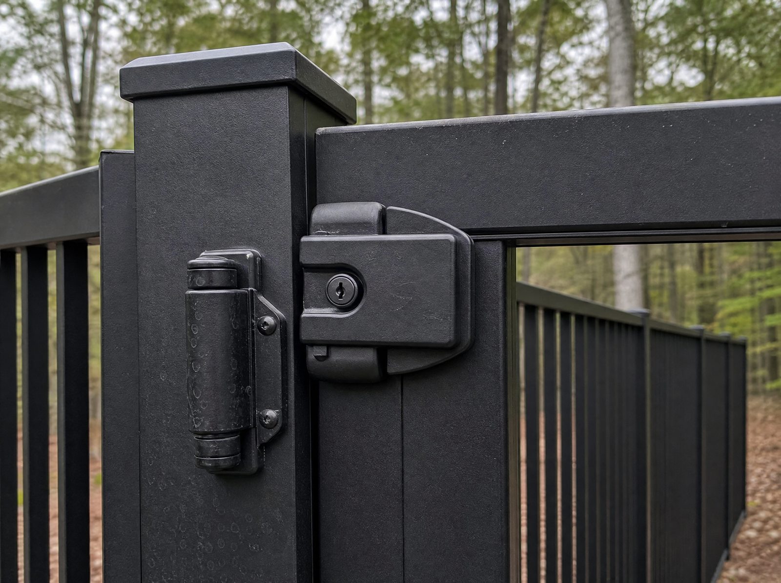 Close shot of a pool gate self-closing hinge and self-latching latch mounted high on the inside of a black aluminum enclosure, latch engaged and gate closed