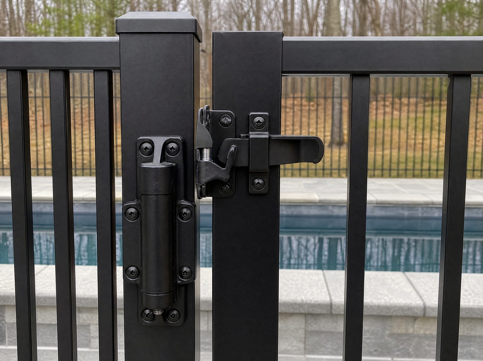 Close view of a black aluminum pool gate's self-closing hinge and self-latching latch mounted high on the inside face with the latch engaged and the gate closed