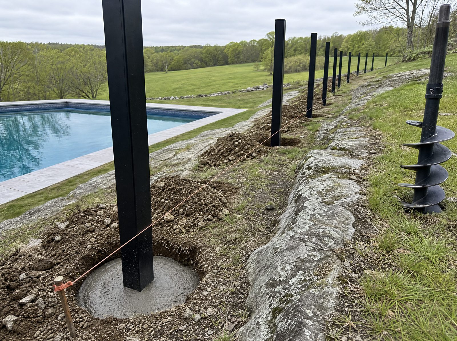 Black aluminum fence posts set plumb in freshly augered holes stepping along a sloped rocky Southbury backyard beside an in-ground pool with no panels hung yet