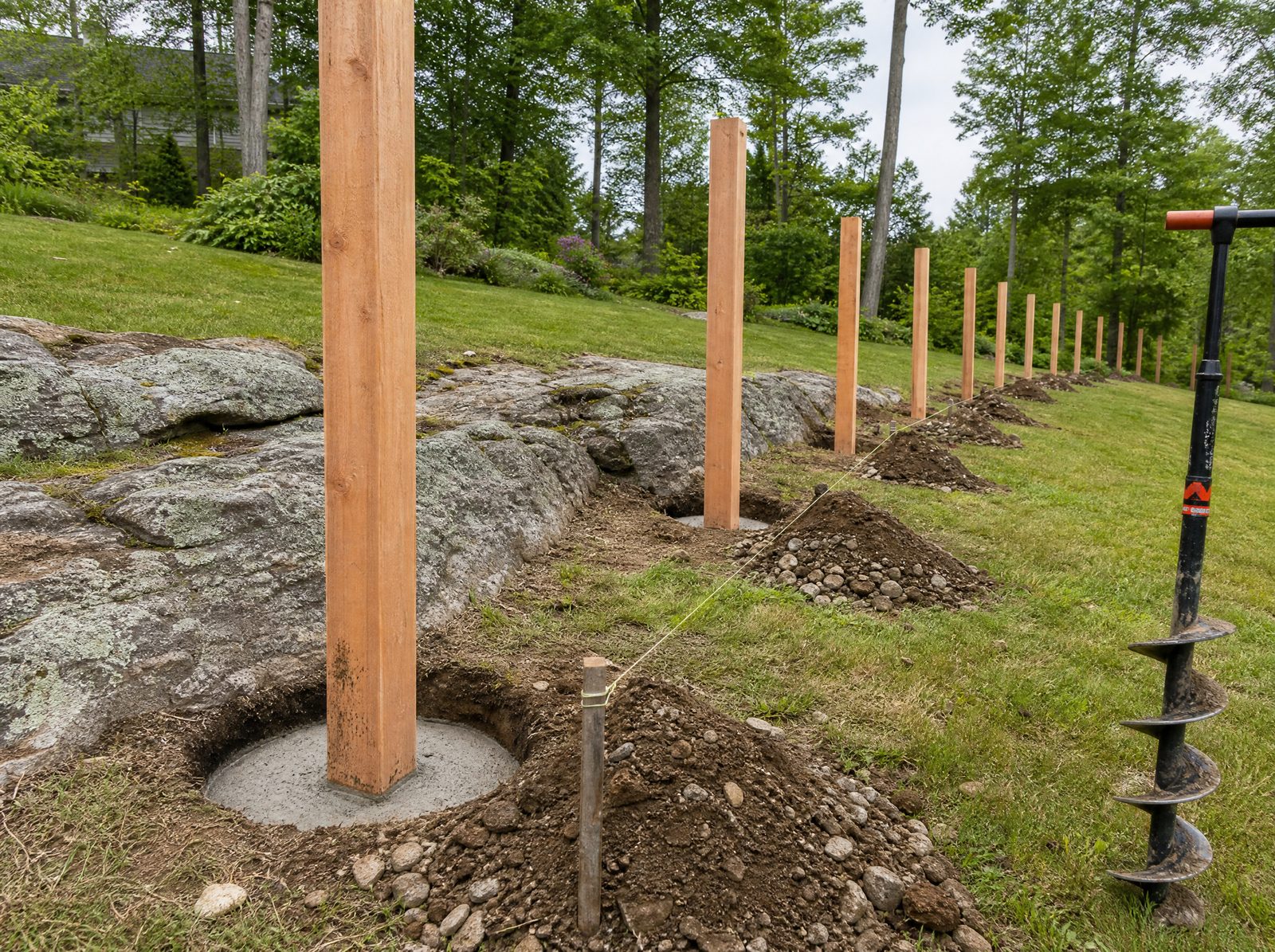 Cedar wood fence posts standing plumb in freshly augered holes along a sloped New Fairfield property line where grey bedrock ledge meets open lawn, with no panels hung yet