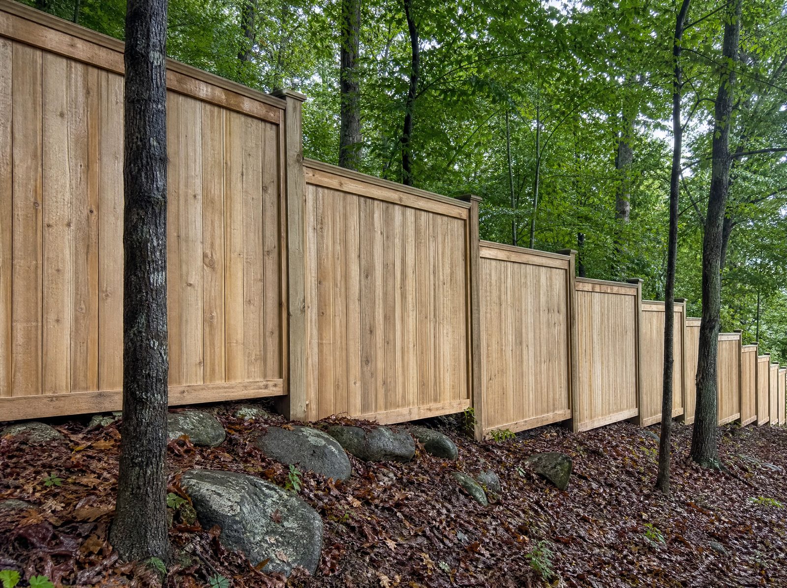 Six foot solid wood board privacy fence stepping down a sloped wooded backyard edge in level rectangular sections with a step offset at each post