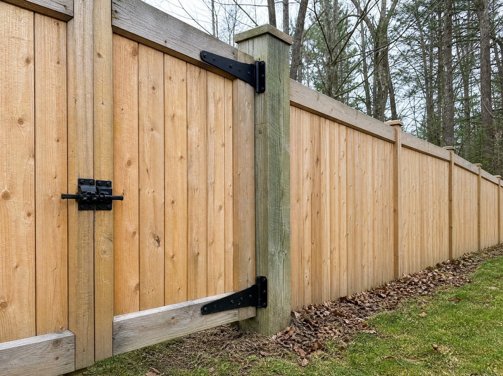 Close honest view of a six foot wood privacy fence gate showing its heavy hinges, latch, and oversized wood gate post with the matching fence continuing away along the line