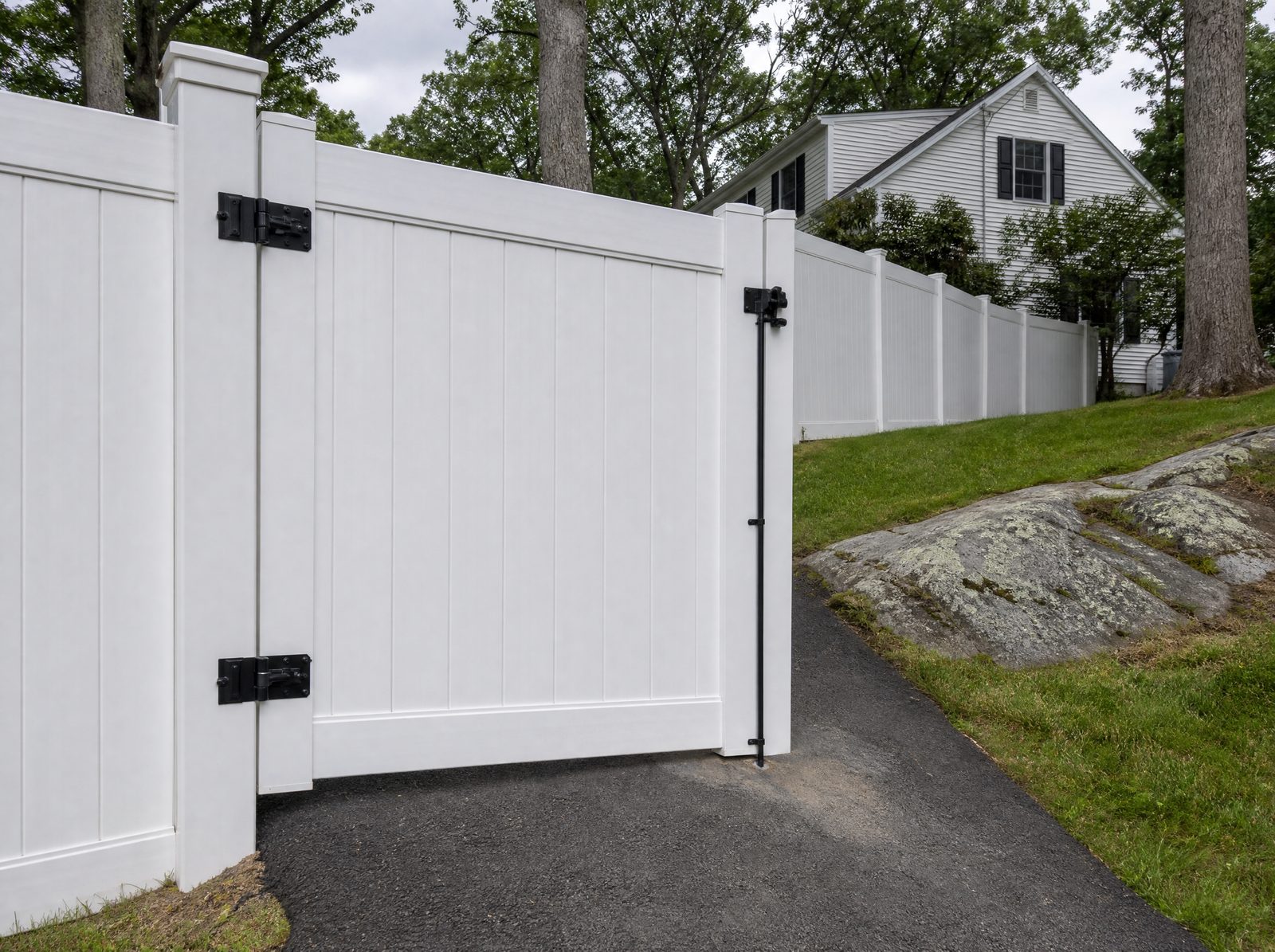 Closed reinforced white vinyl gate hung on a heavy white vinyl post beside a sloped suburban driveway, with matching six foot white vinyl privacy fence continuing away from it