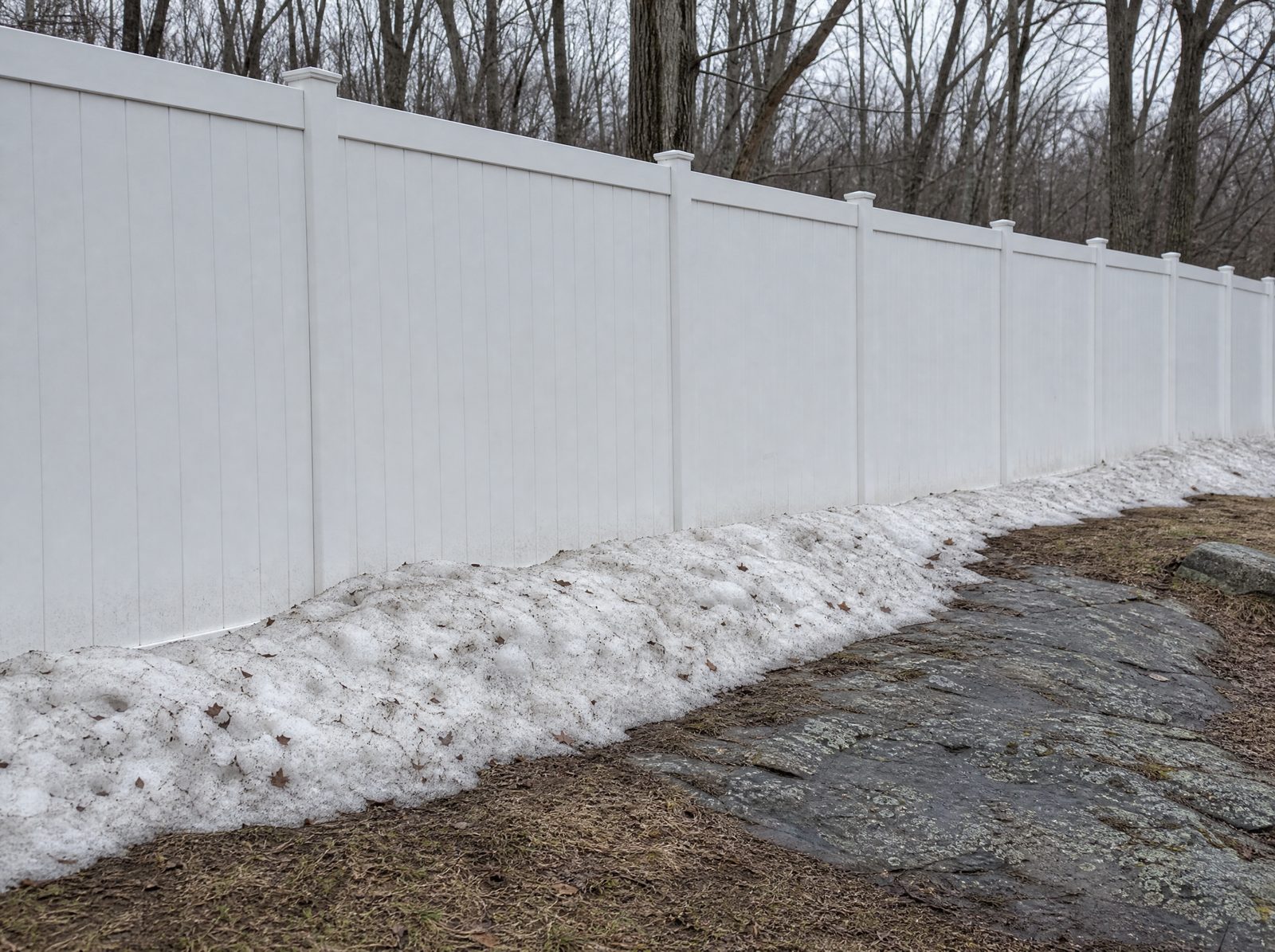 Six foot white vinyl privacy fence in late winter with a light crusted snow drift banked against the base on the windward side and bare hardwoods behind