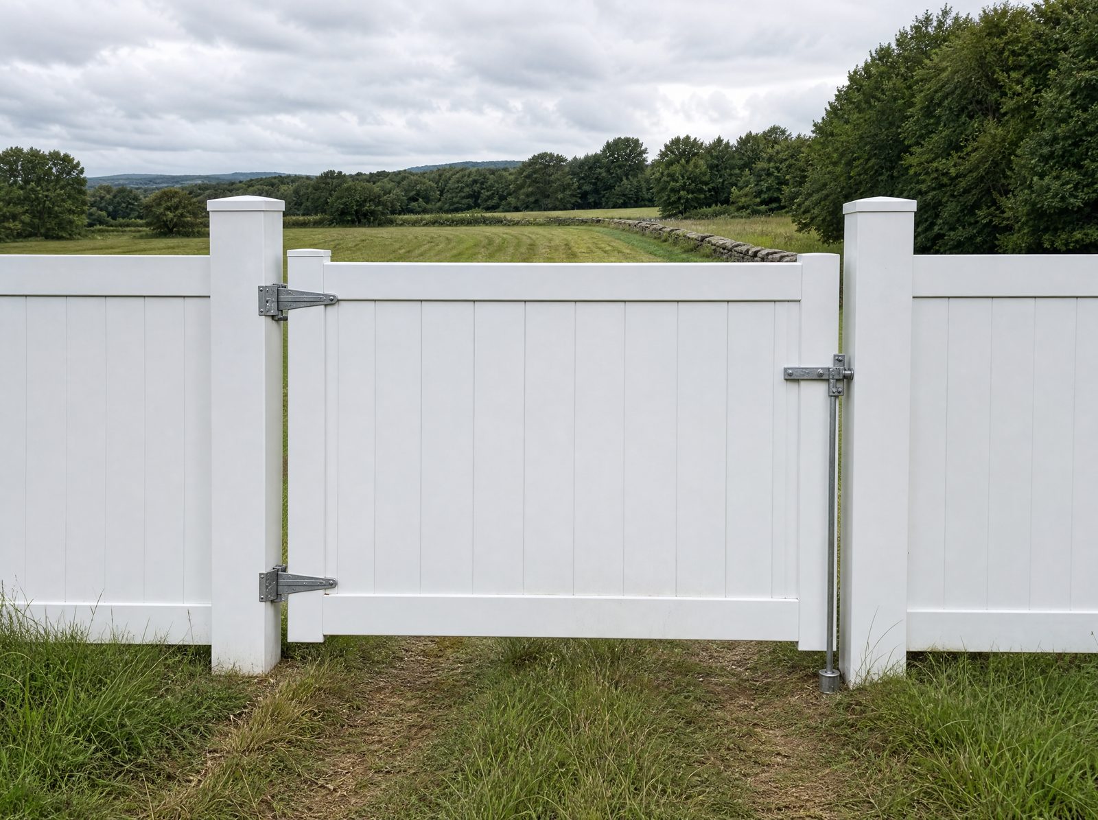Reinforced white vinyl gate closed across a mowed farm track through a hay field, hung on a heavy reinforced post with visible heavy hinges and a drop rod, with the matching six foot white vinyl fence continuing along the field boundary