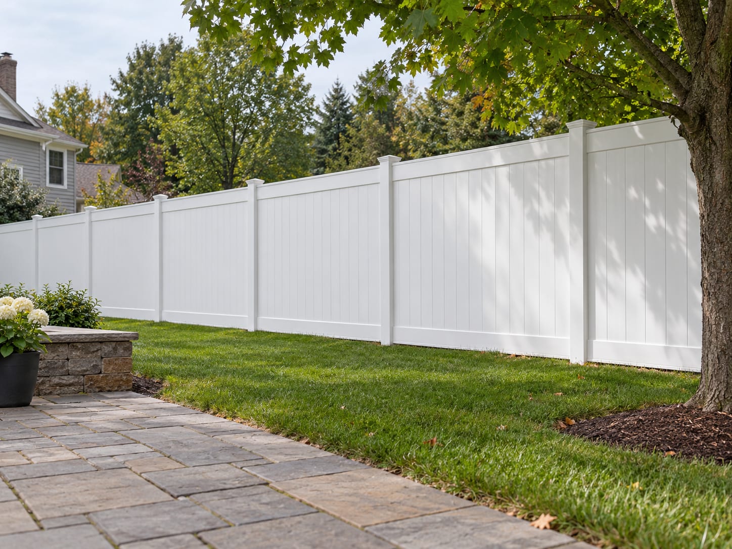 A white vinyl privacy fence runs straight along the side of a level backyard in western Connecticut, with a small patio and a maple tree on the near side.