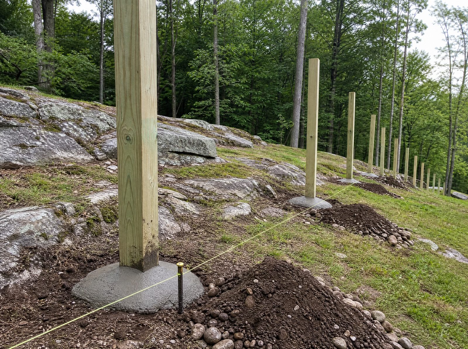 Pressure-treated wood fence posts set plumb along a sloped rocky property line with concrete crowned and sloped away from each post and no panels hung yet