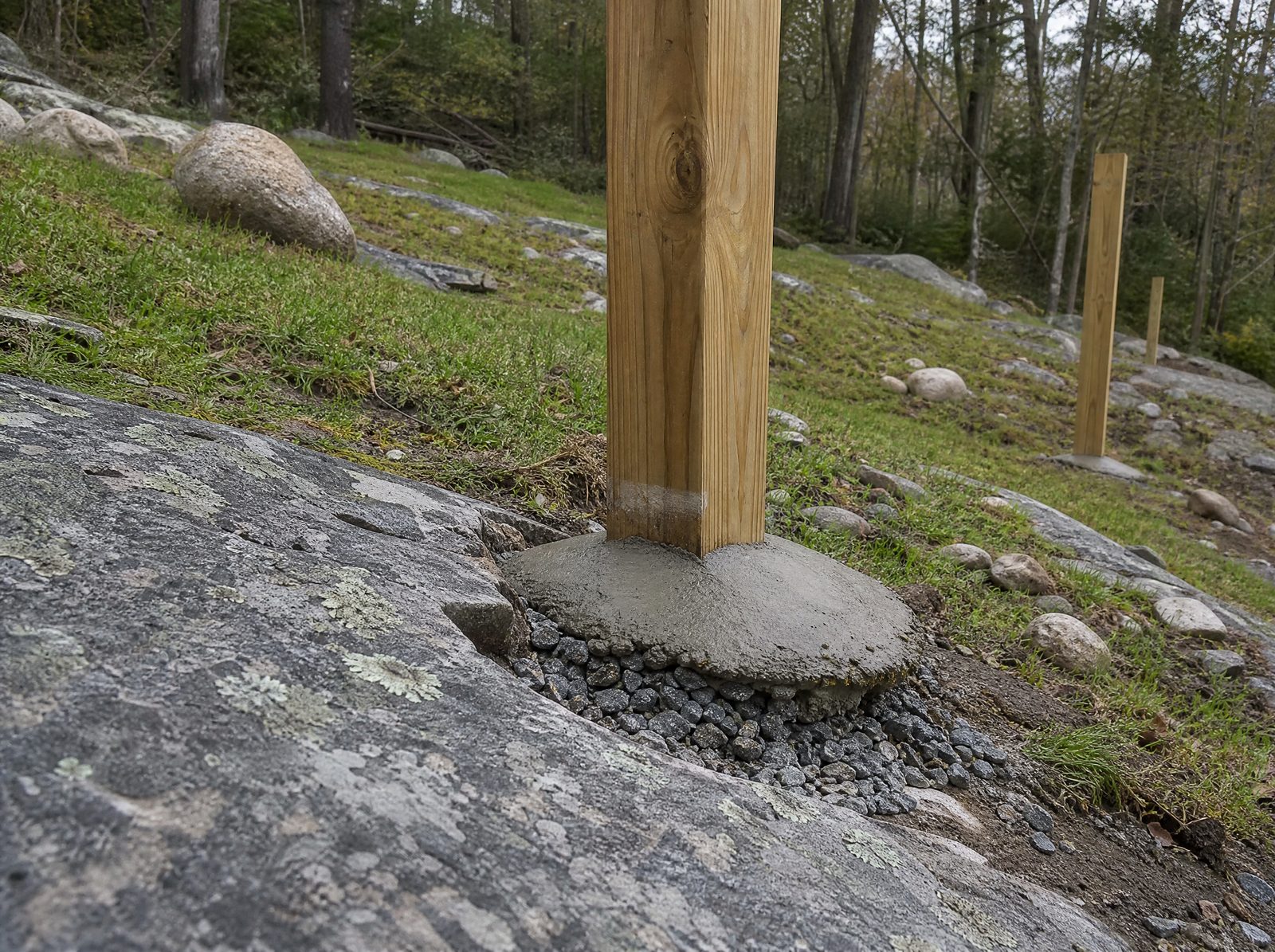A single wood fence post set with drainage at its base, crowned concrete and gravel shedding water away, on a rocky sloped Sherman lot