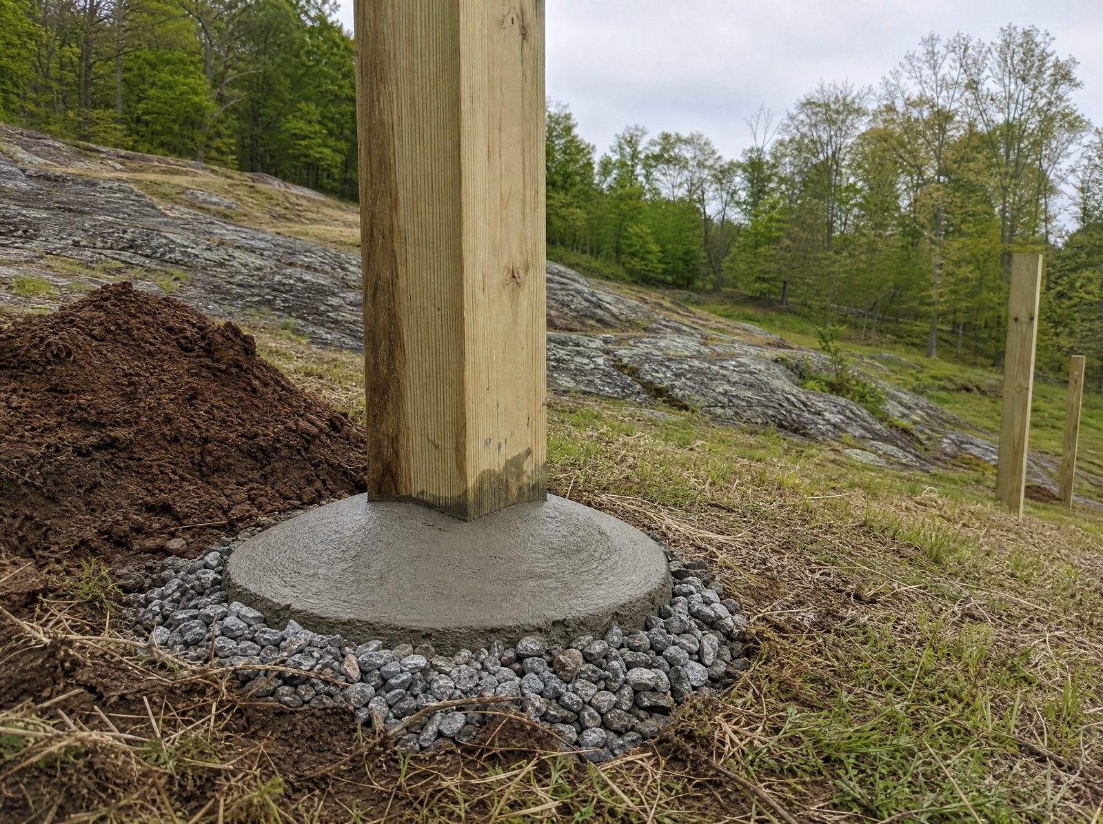 Close low view of a single wood fence post set with crowned concrete sloped away from the wood and gravel around the collar on a rocky sloping lot