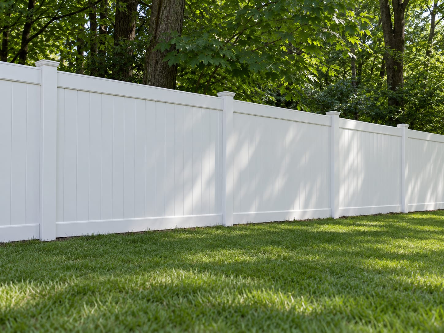 A white vinyl privacy fence runs straight along the edge of a shaded backyard, with a clean lawn in front and maple trees behind.