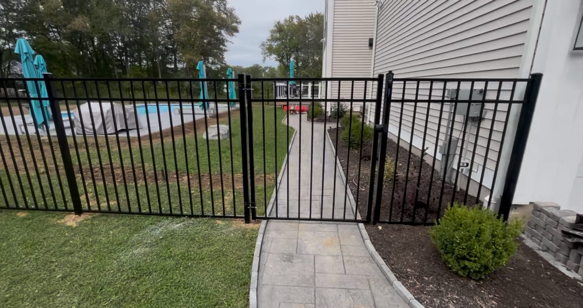 Black aluminum fence with a single walk gate across a paver walkway beside a house, pool visible in the background