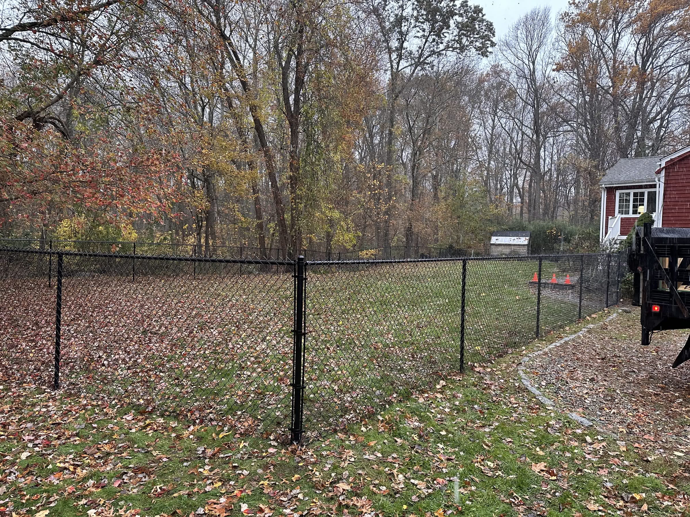 Black chain-link fence enclosing a leaf-covered backyard beside a red house in autumn