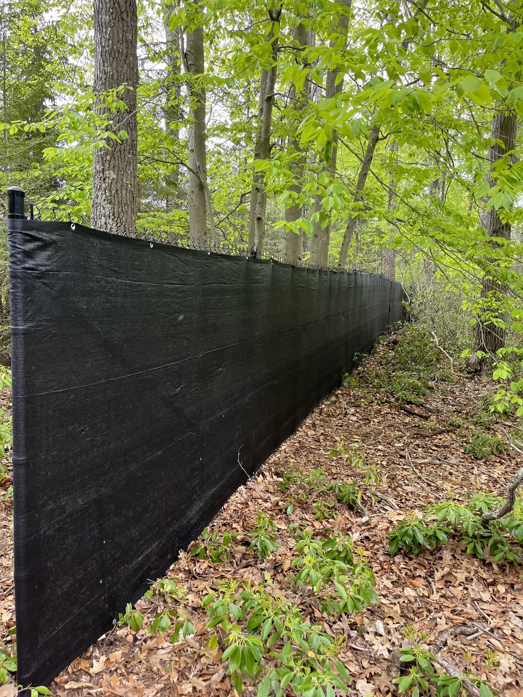 Black privacy screen mesh attached to a chain-link fence running through a wooded area