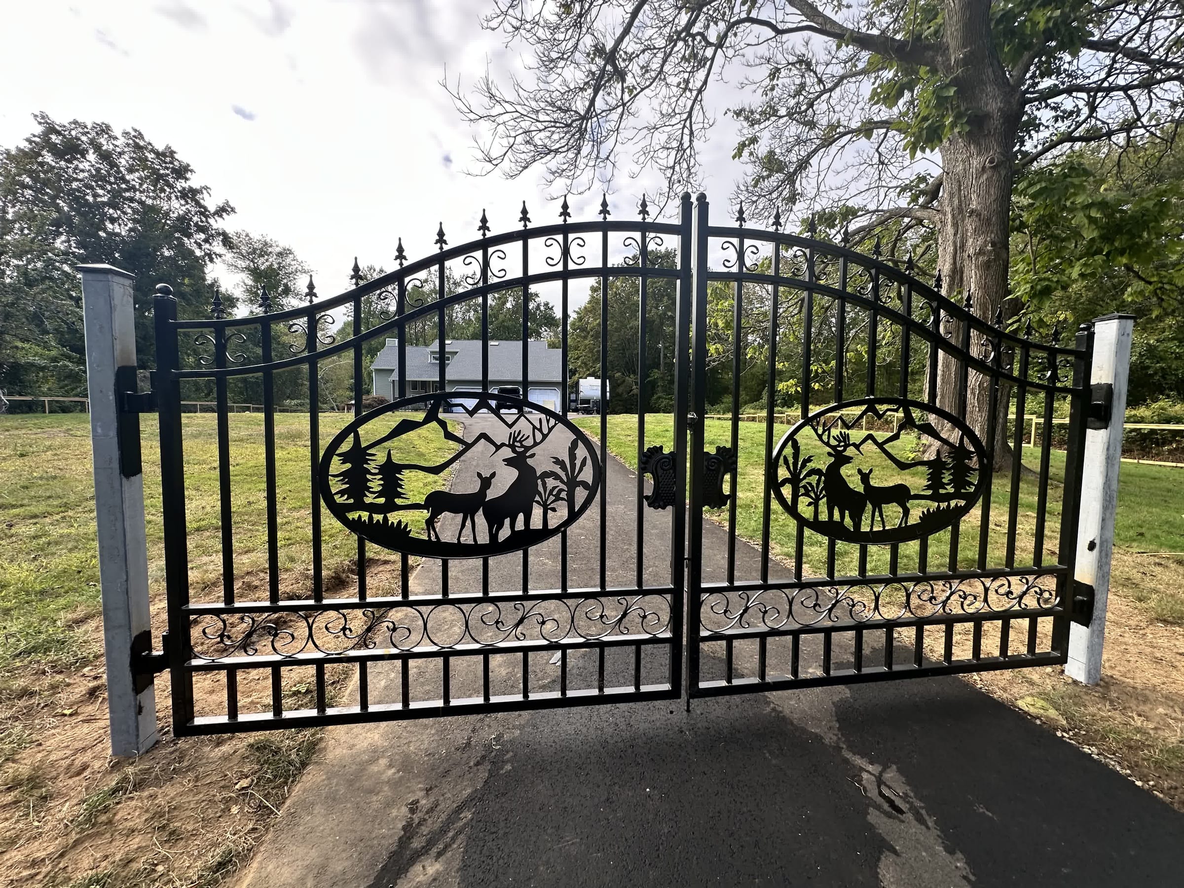 Black ornamental iron double driveway gate with scrollwork and deer-and-mountain medallions between metal posts