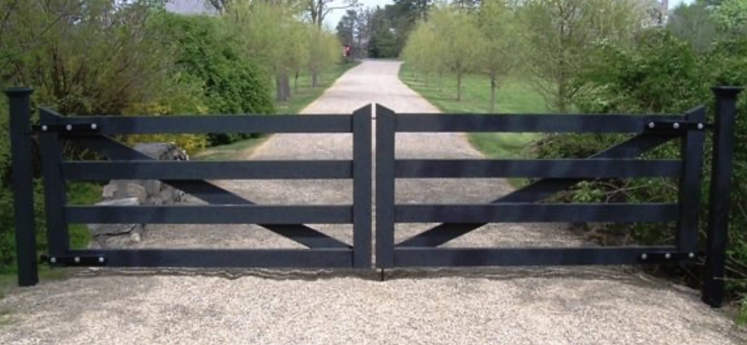 Black five-bar wood estate gate with X-bracing across gravel driveway lined with trees