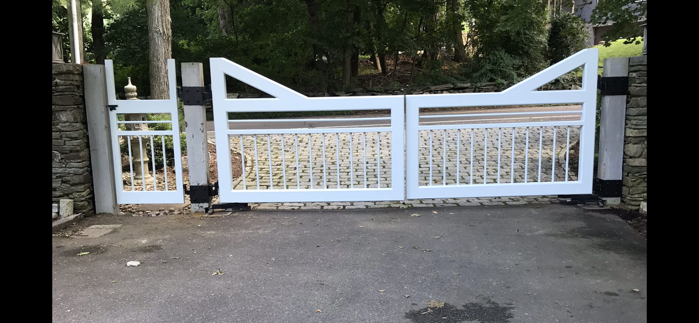 Black four-bar wood estate gate with X-bracing across gravel driveway lined with willow trees