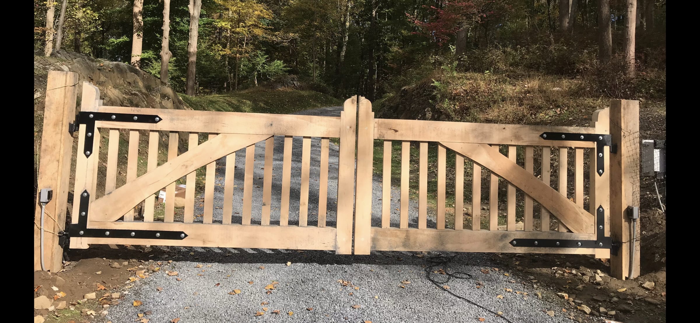 Natural unfinished wood double driveway gate with black strap hinges and Z-bracing on gravel drive in wooded setting