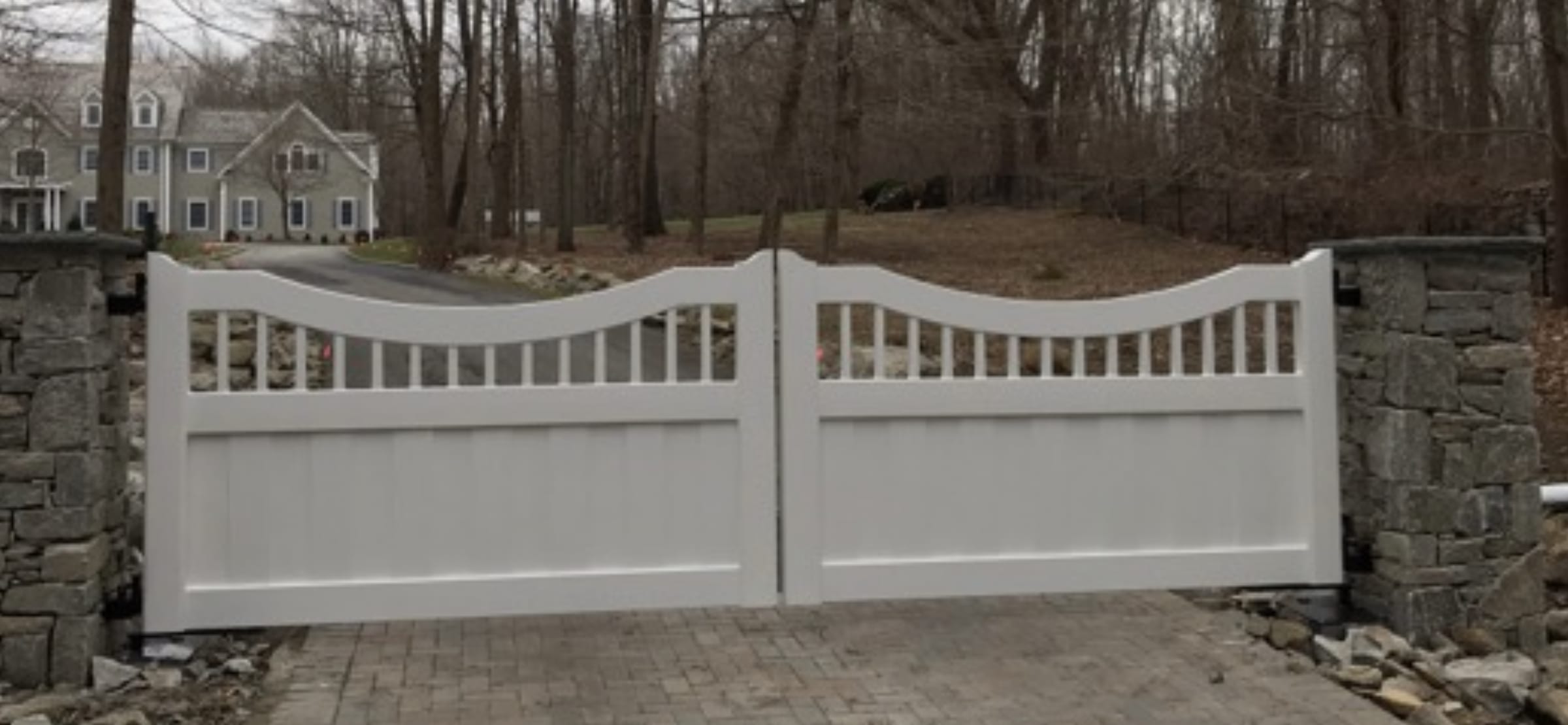 White arched double driveway gate with spindle top and solid lower panels between stone pillars on paver drive