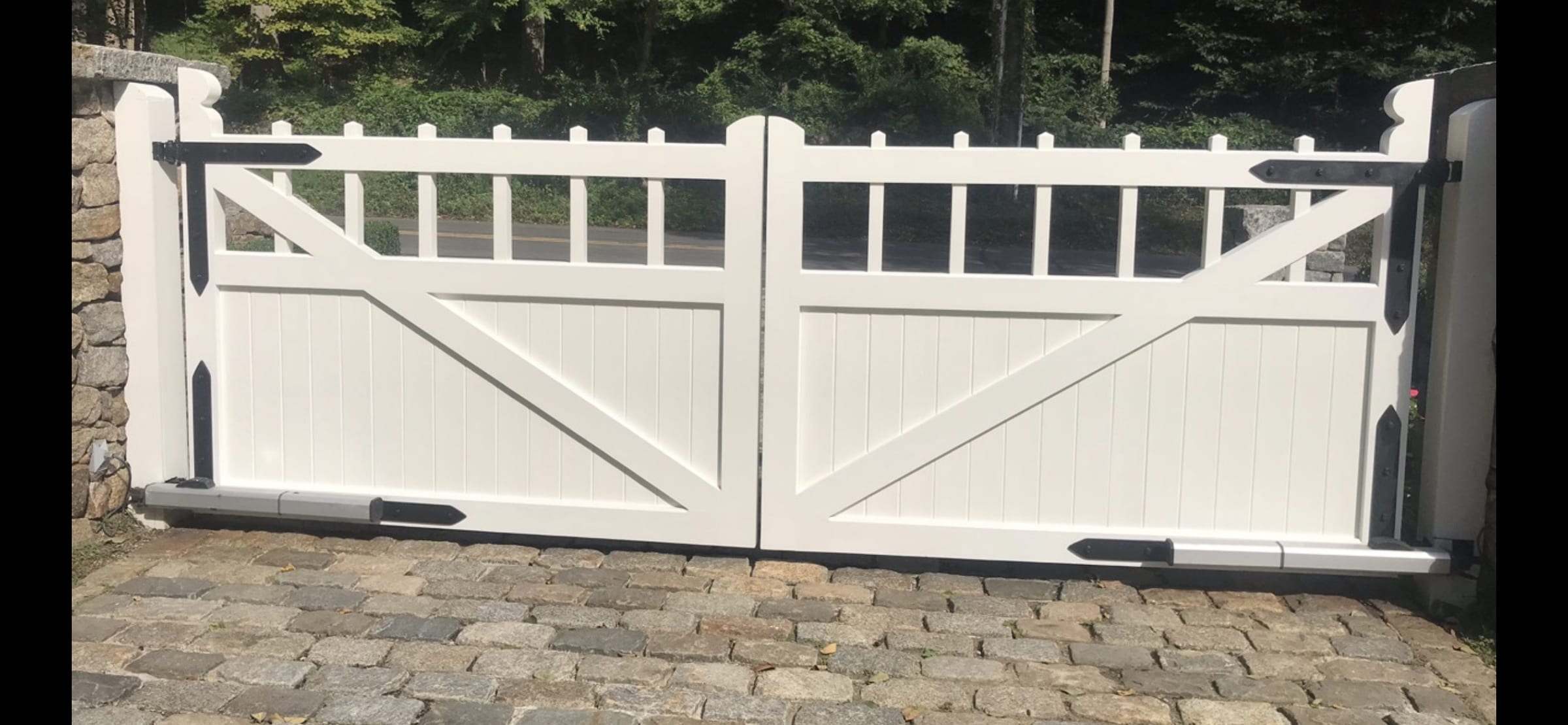 White board-and-batten double driveway gate with picket top and Z-bracing between stone walls on cobblestone drive