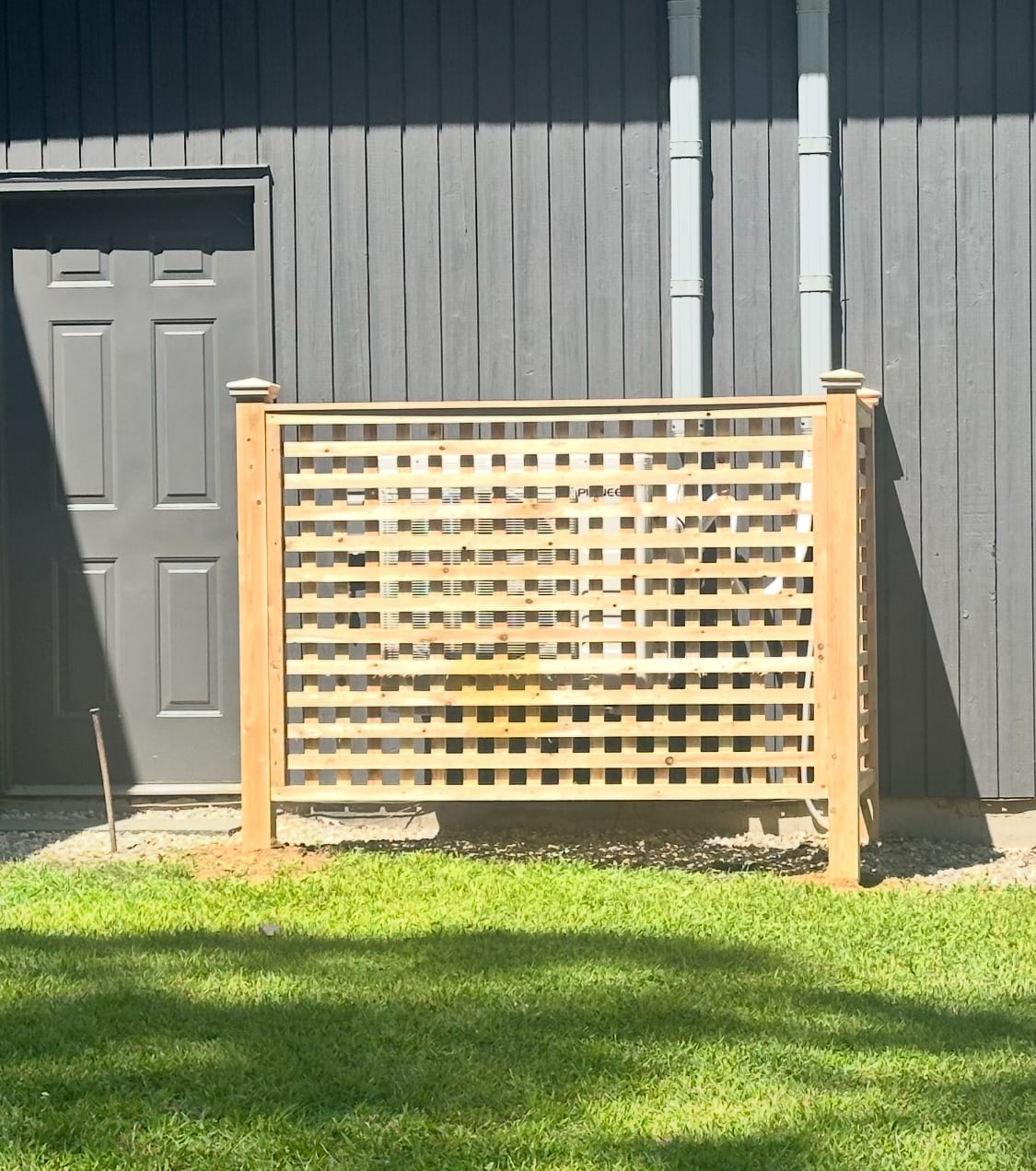 Freestanding natural cedar lattice privacy screen mounted on two posts in a grass yard beside a dark gray building.