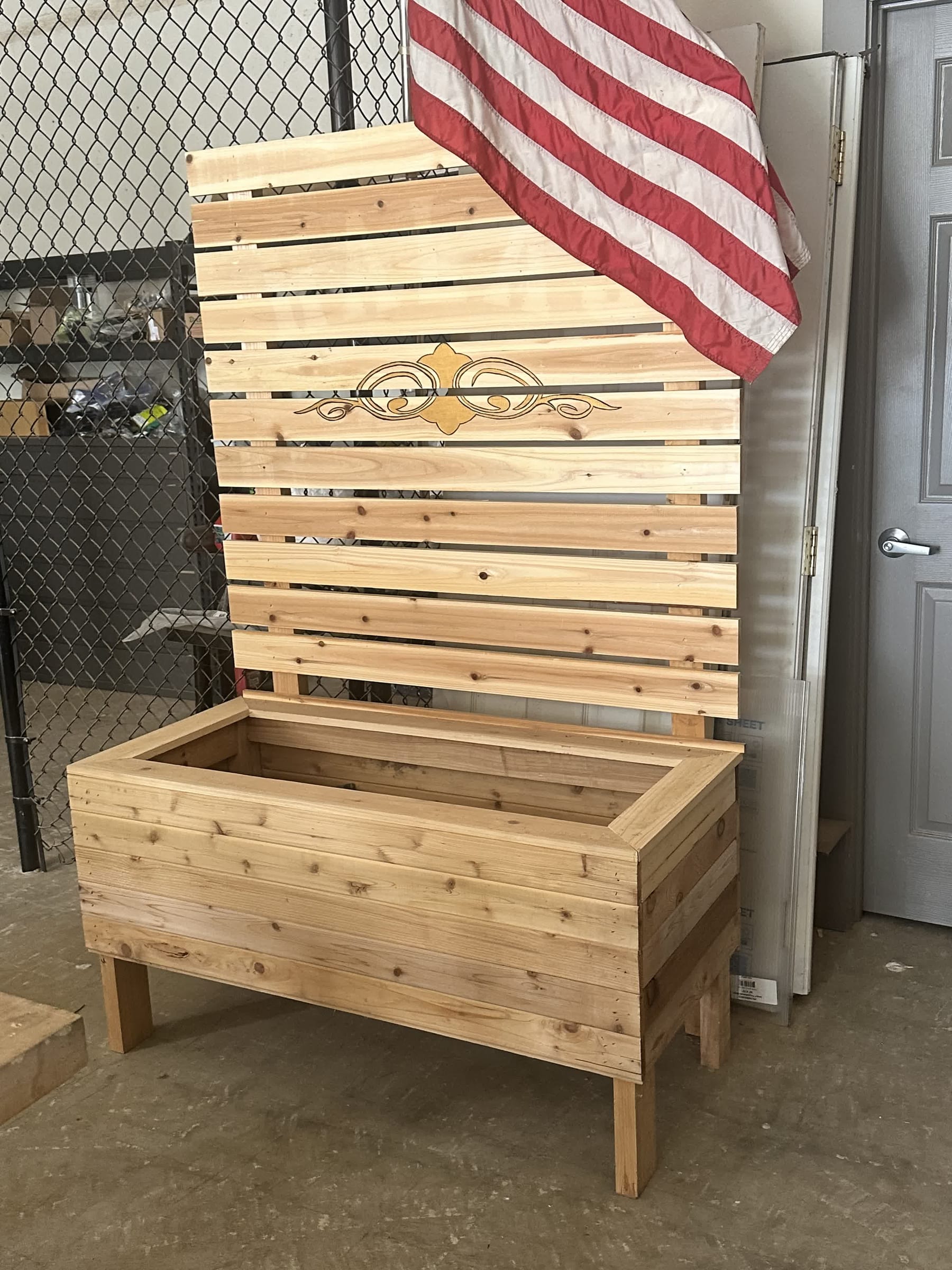 Cedar raised planter box with attached horizontal-slat privacy screen and decorative scrollwork engraving, photographed in a workshop.