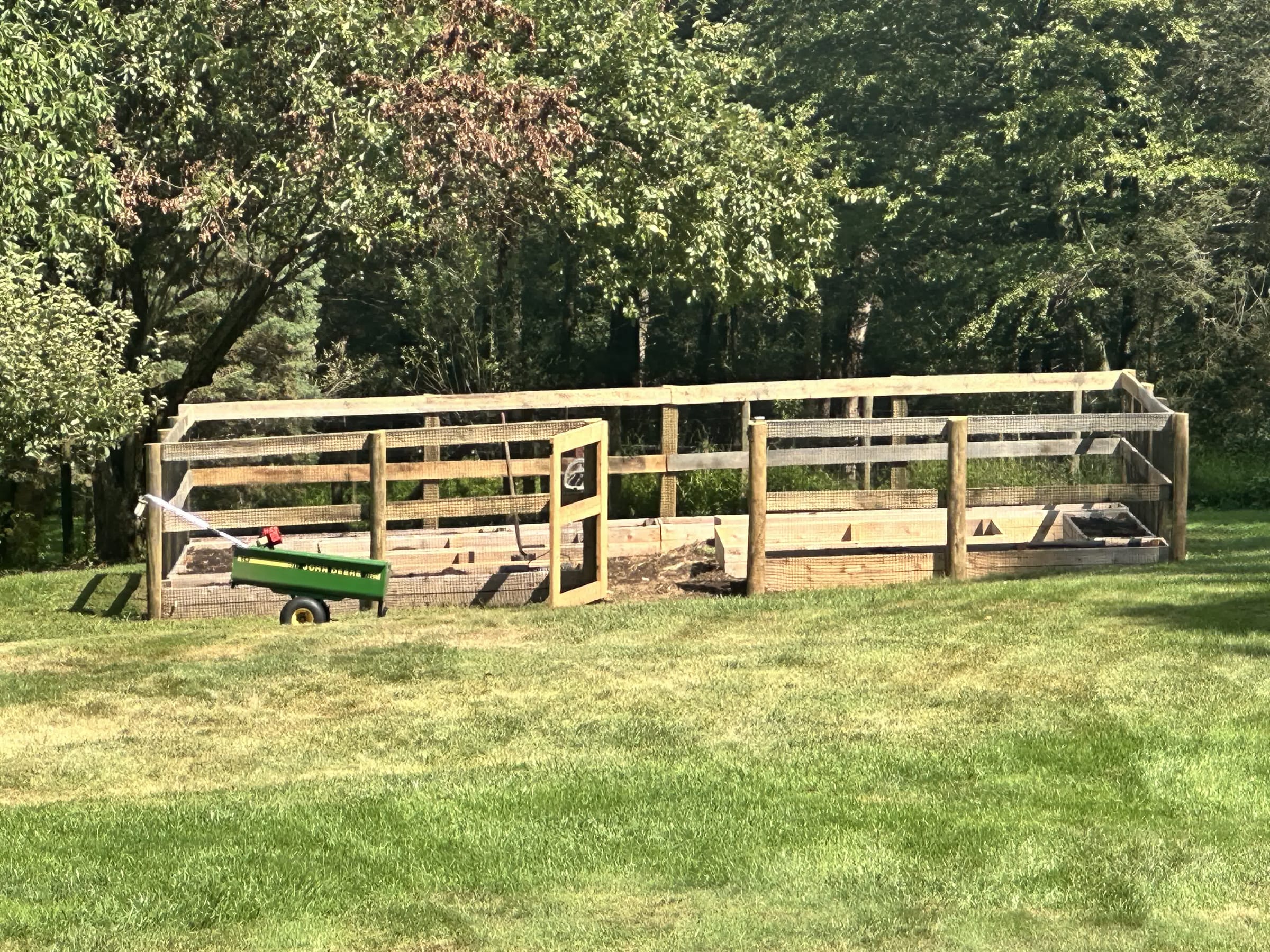 Long wood post-and-board garden enclosure with wire mesh and a gate at a wooded lawn edge; toy John Deere wagon nearby.