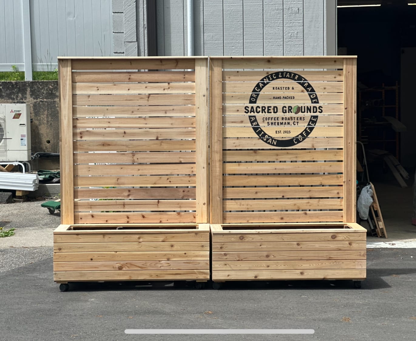 Two cedar raised planter boxes on casters with slat privacy screens, one screen laser-engraved with a Sacred Grounds Coffee Roasters logo.