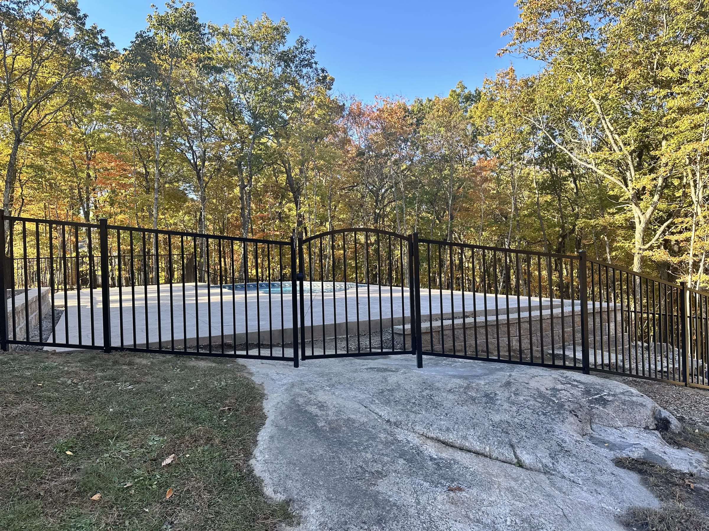 Black aluminum fence with a double arched gate around a newly built inground pool and concrete deck in wooded surroundings